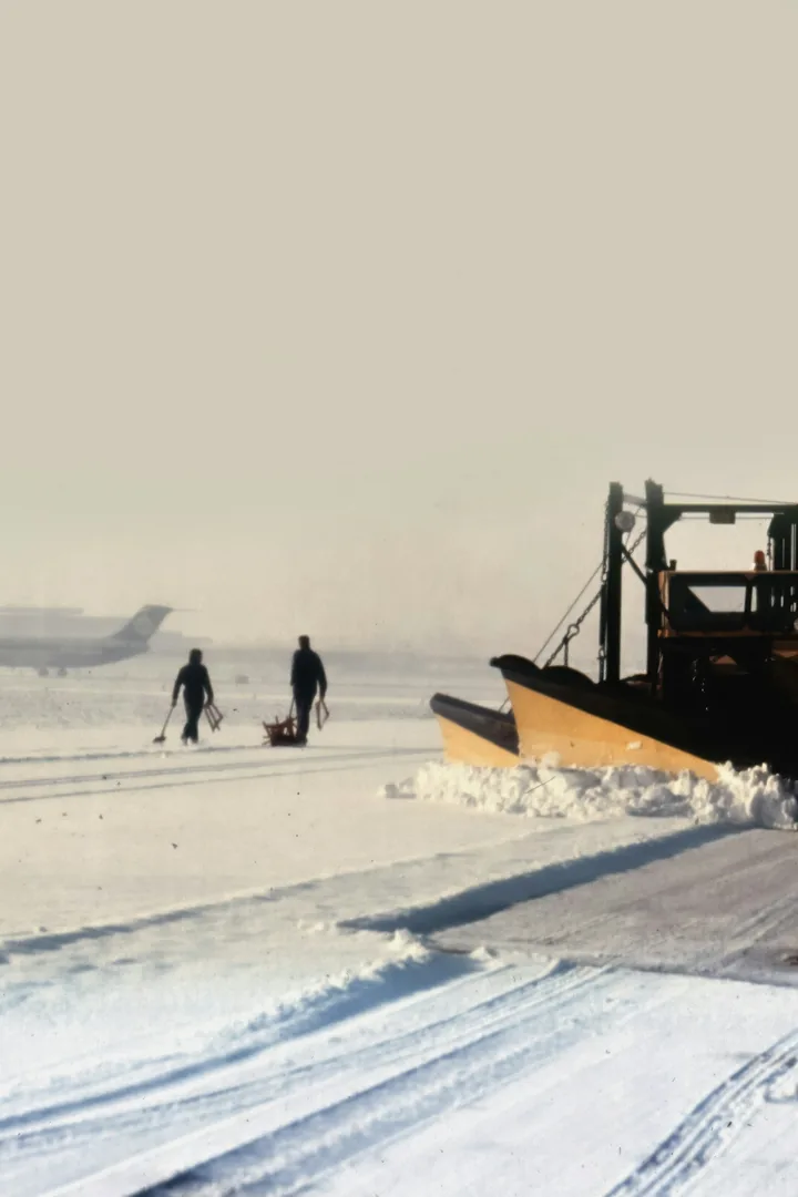 A snow plough clearing a runway, two people walking beside it